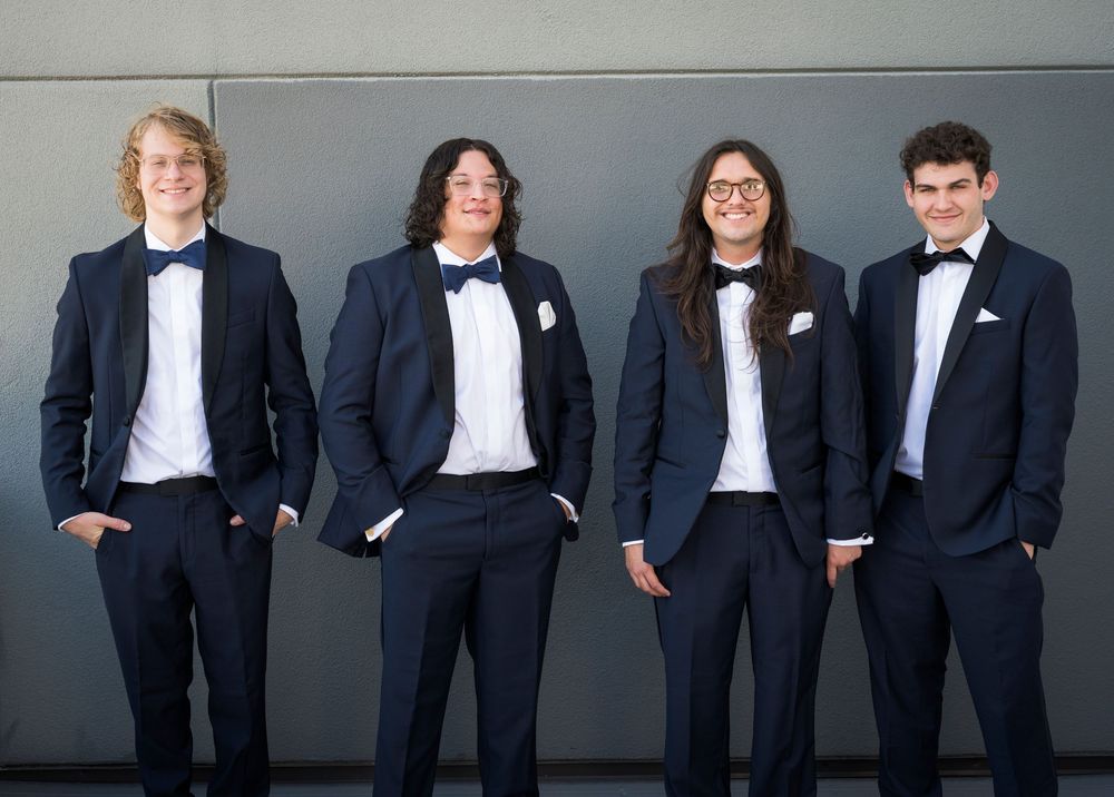 Four groomsmen in matching navy tuxedos with bow ties posing together against a gray wall.