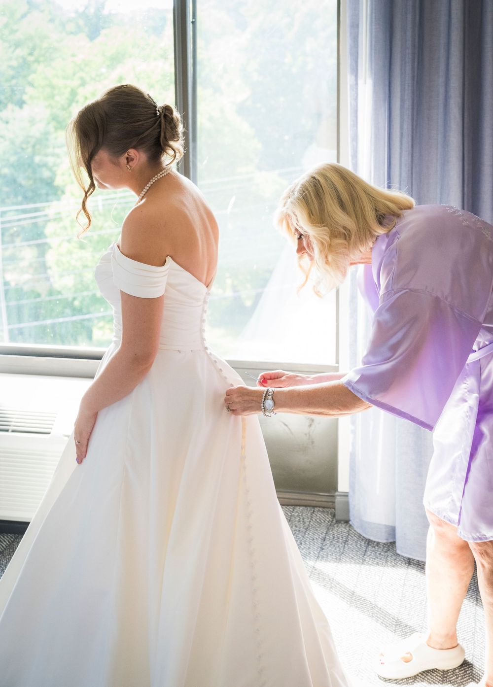 A bride in an off-shoulder white gown gets her dress buttoned by a woman in lavender near a bright window.