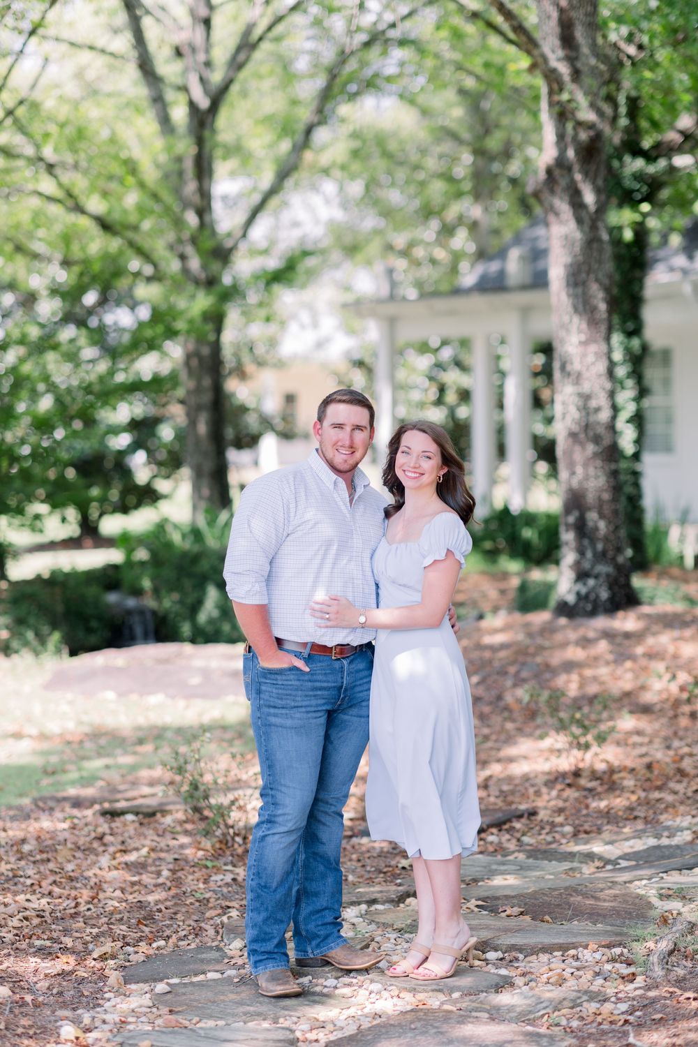 A couple in light-colored clothing poses together on a tree-lined path in front of a white building on a sunny day.