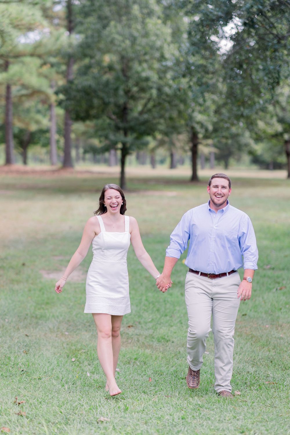 A couple walks and laughs together on a grassy field surrounded by trees on a sunny day.