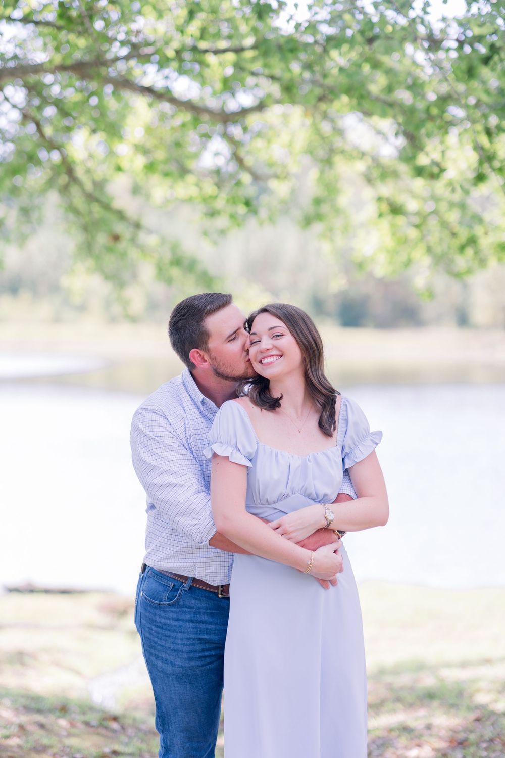 A couple in light blue clothing embraces lovingly outdoors in a natural setting with green trees and water in background.