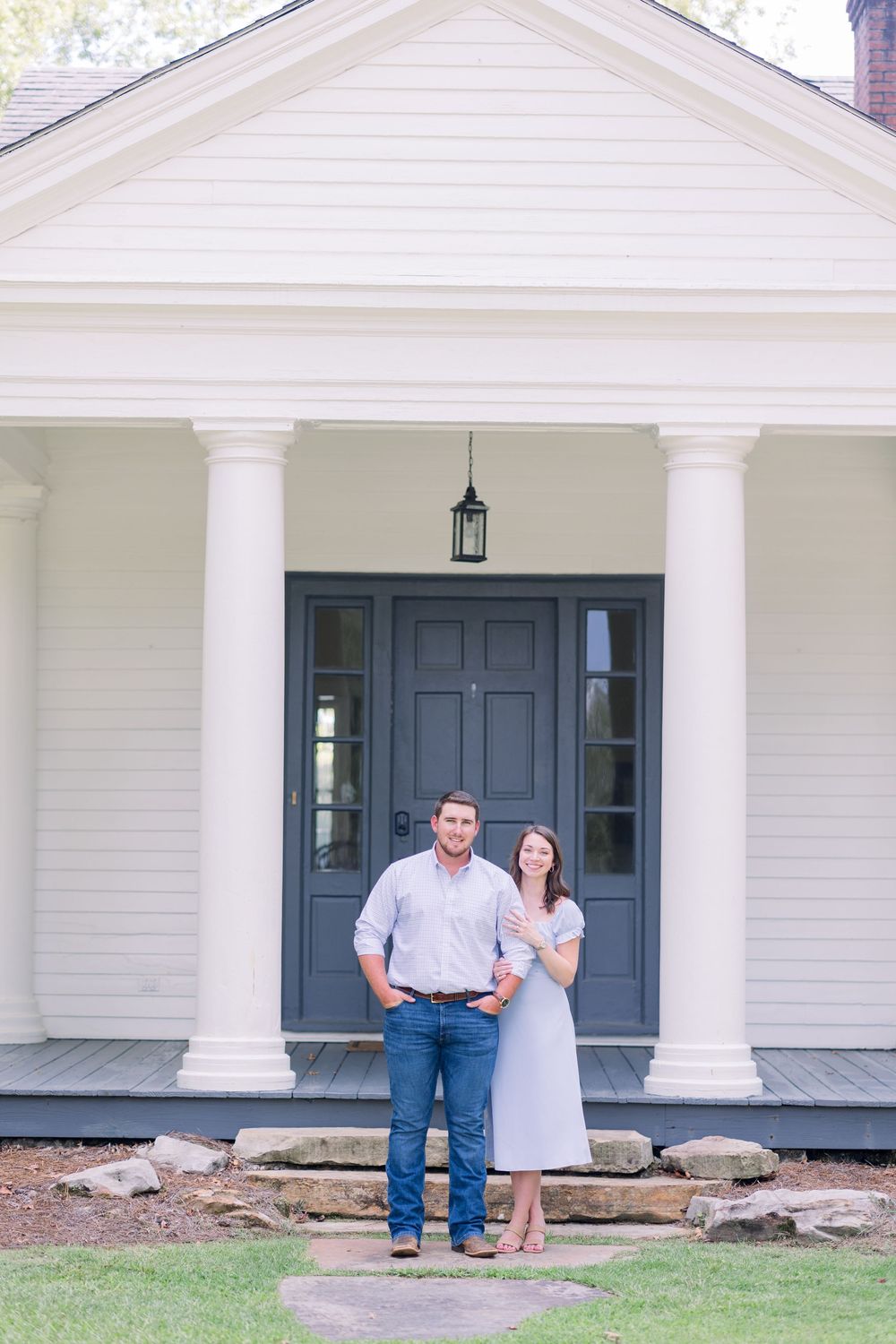 A couple stands on a white porch with classic columns in front of a house with navy blue doors.