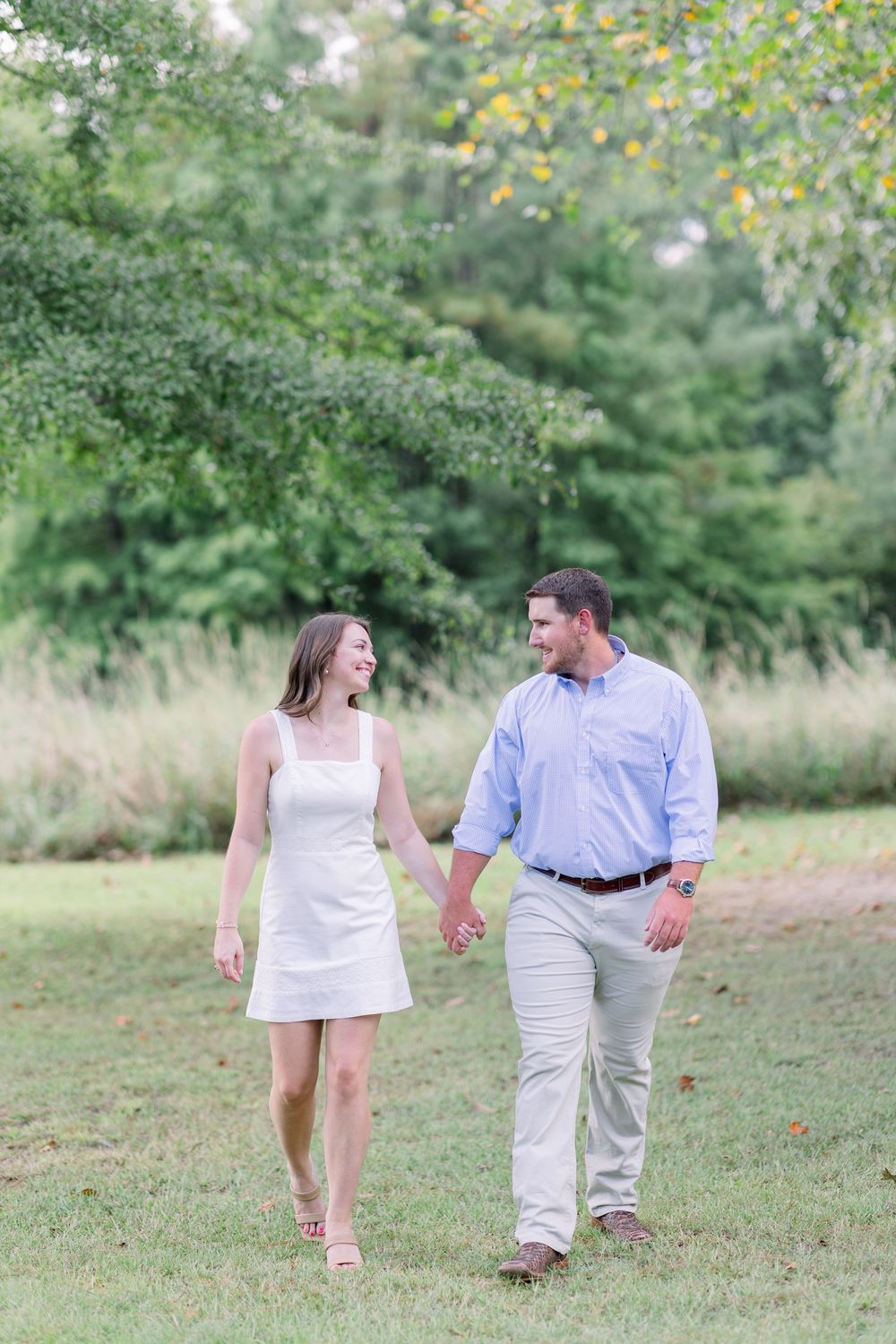 A couple in white and beige attire walks hand in hand through a lush green field on a sunny afternoon.