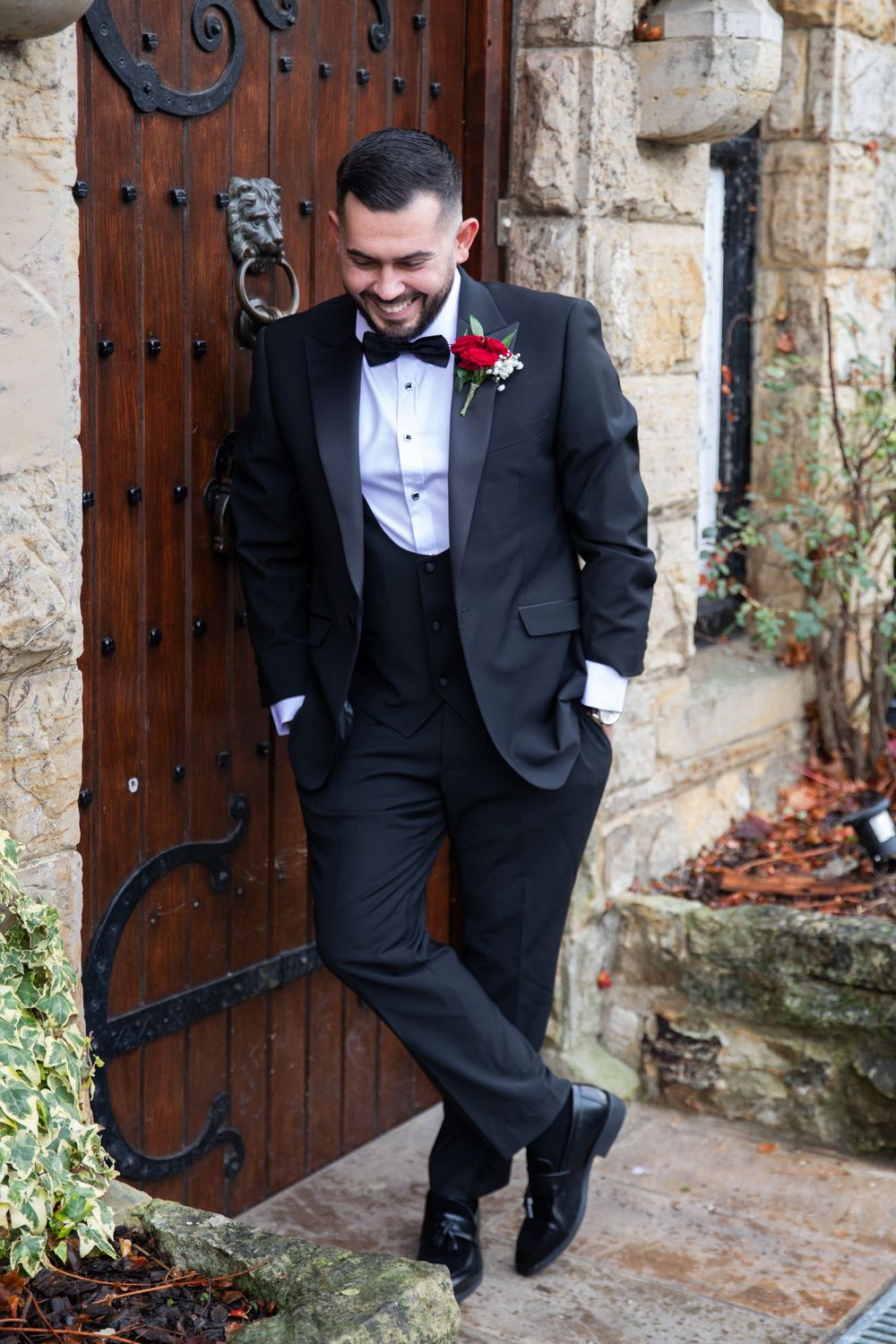 Groom in black tuxedo and bow tie poses playfully by rustic wooden door and stone wall with red boutonniere.