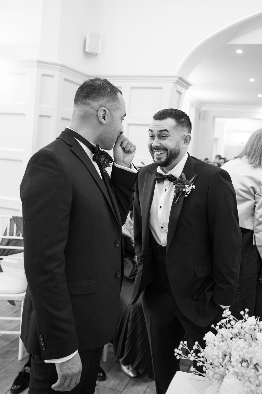 Wedding party members in black tuxedos share a joyful candid moment during formal photos.