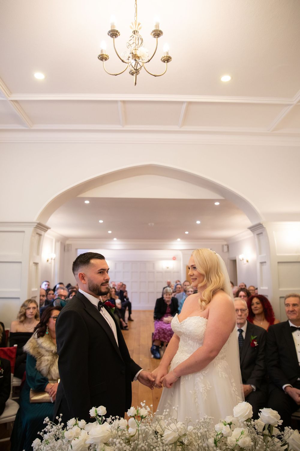 Wedding ceremony taking place in an elegant white ballroom with chandelier and floral decorations.
