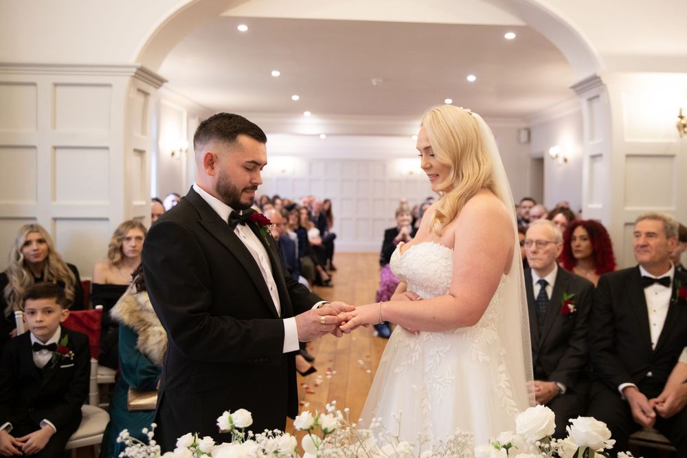 Elegant wedding reception in a bright white ballroom with guests seated at round tables decorated with floral centerpieces.