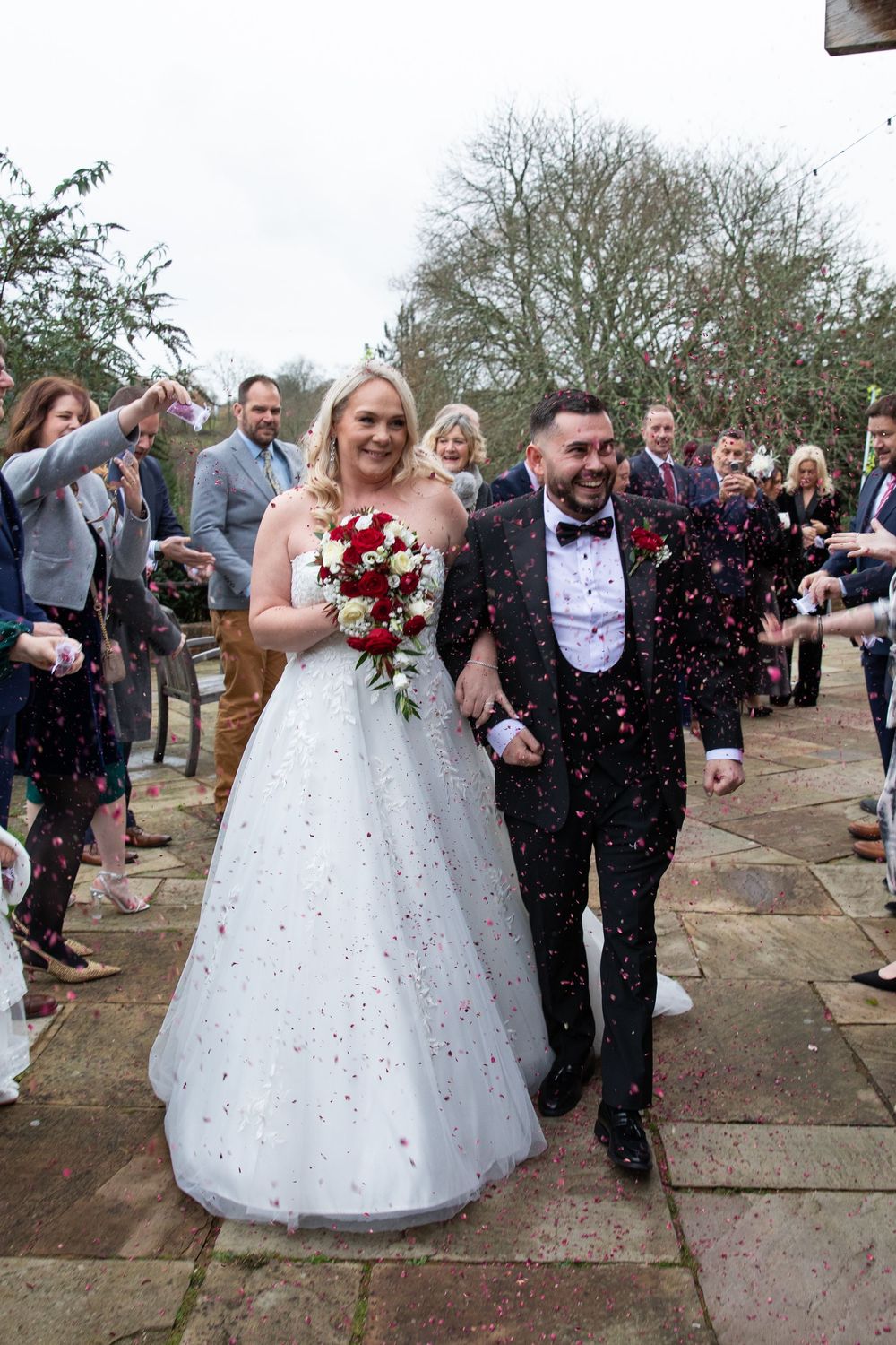 Newlyweds walk through shower of red confetti as guests celebrate outside venue.