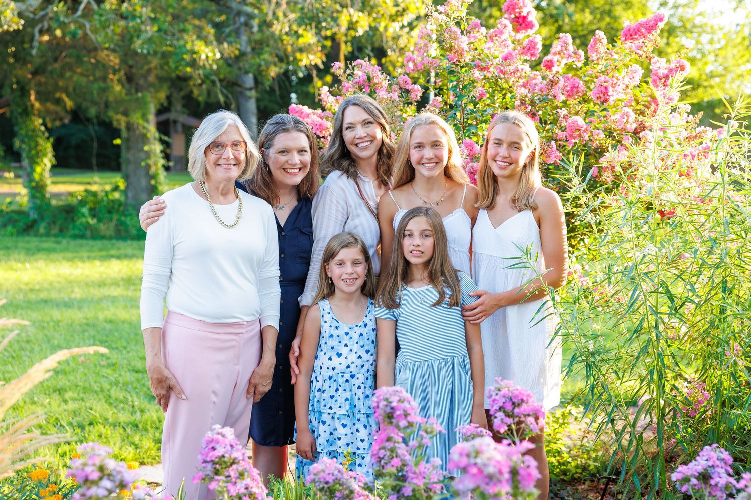 A group of people pose together outdoors in a garden filled with vibrant pink and yellow flowers on a sunny day.