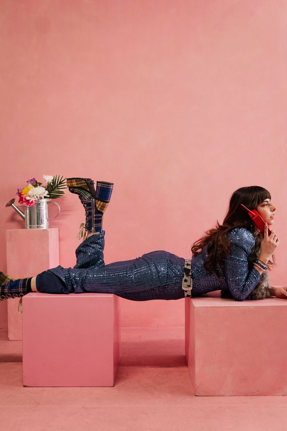 Person lying on pink blocks wearing denim jacket and jeans with flowers in a vase against pink background.