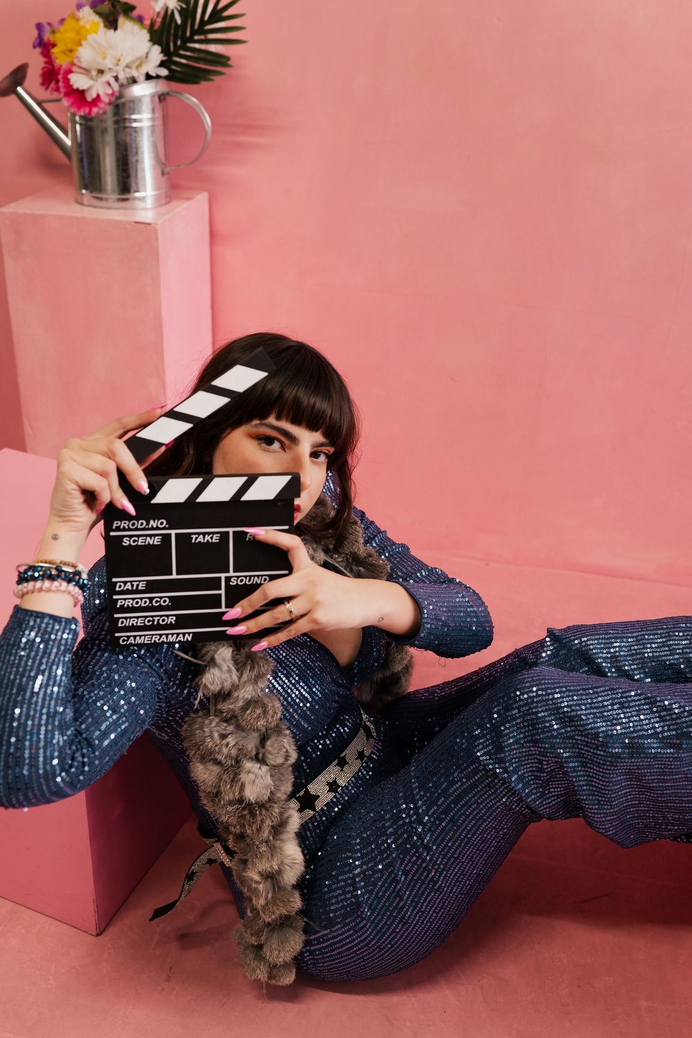 Movie clapperboard held against pink background with sparkly blue dress and decorative palm fronds in styled photoshoot.