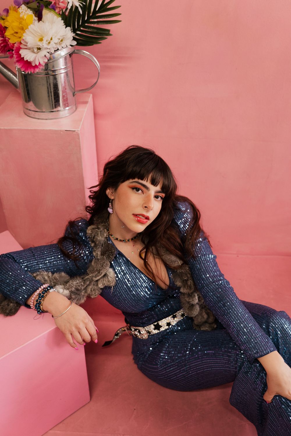 Fashion model in sparkly navy blue jumpsuit poses on pink backdrop with dramatic lighting and styling.