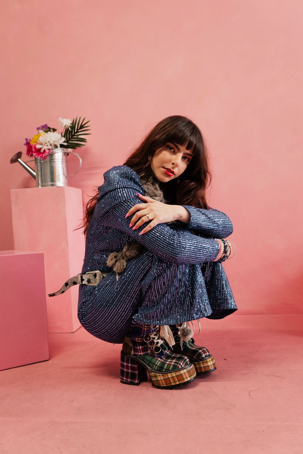 Person in blue sweater and plaid skirt curled up on pink floor with potted plants in minimalist setting.