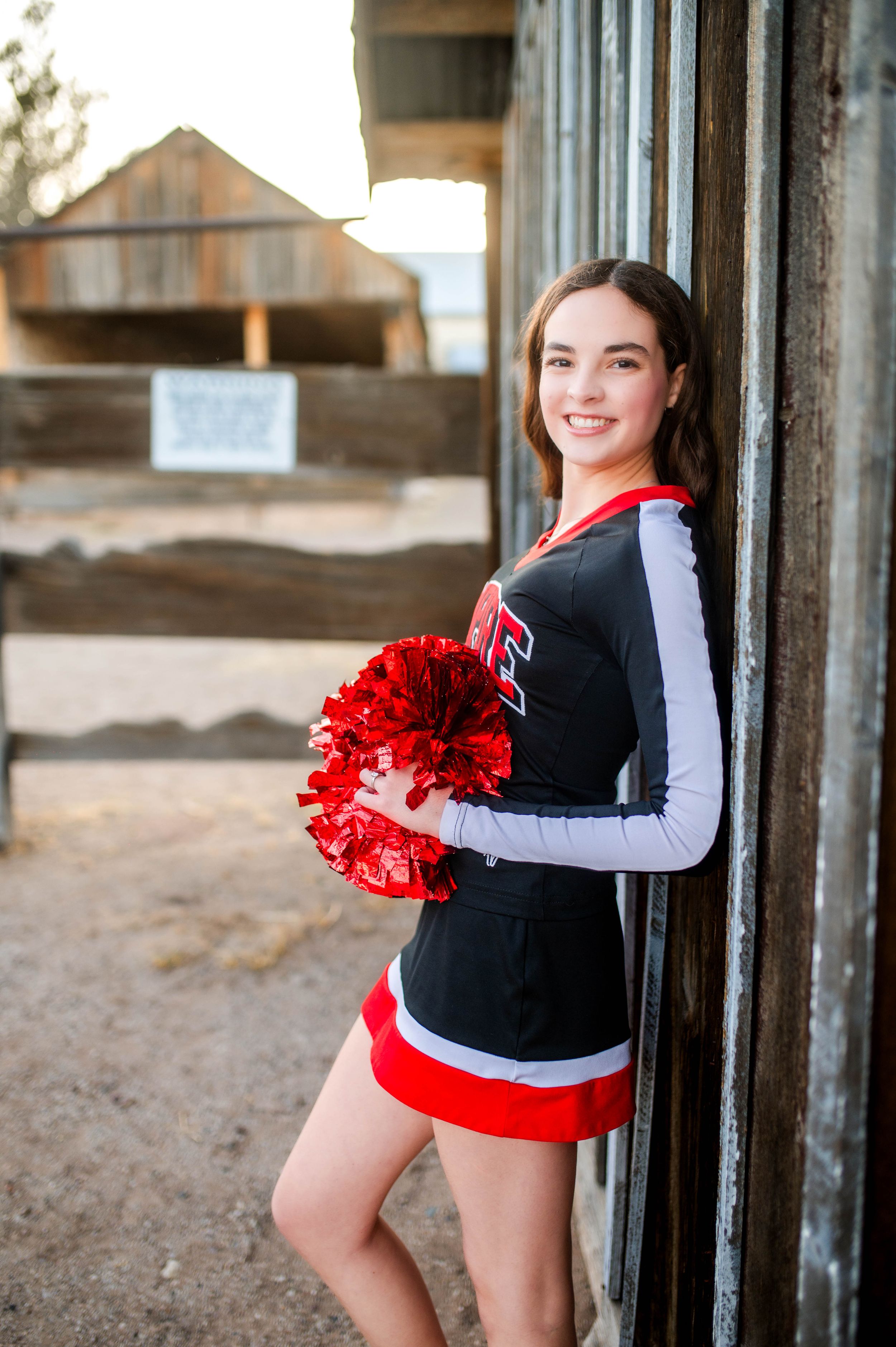 Cheerleader in black and red uniform holds red pom-poms while posing against a rustic wooden wall.