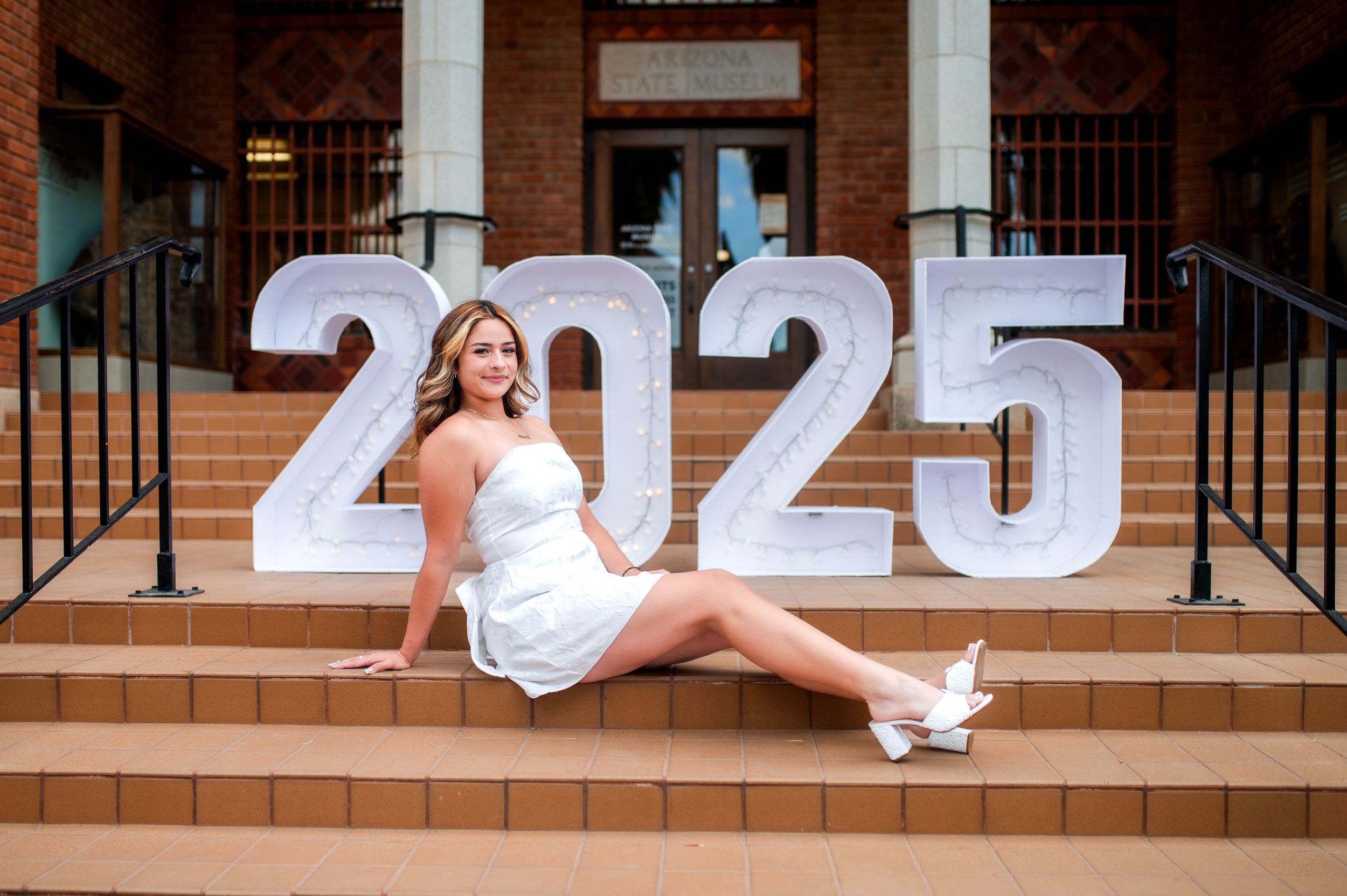 Young woman in white dress posing on brick steps beside illuminated 2025 graduation sign.