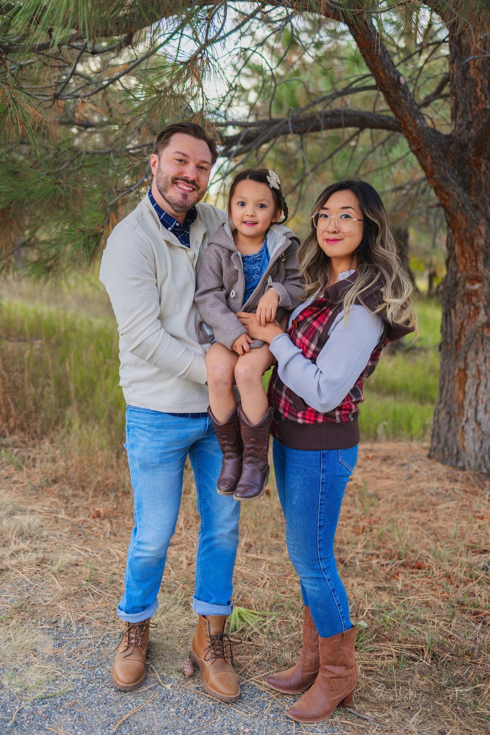 A family poses together outdoors in casual attire among pine trees on a fall day