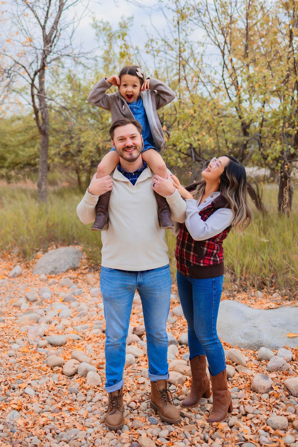 A family shares a joyful moment together outdoors among autumn leaves and rocks in a natural setting.  Little girl sat on mans shoulders.