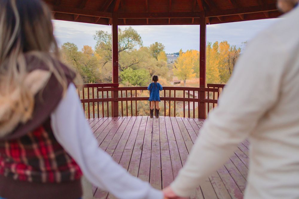 A couple in warm coats stands on a wooden bridge deck surrounded by autumn trees and golden foliage.