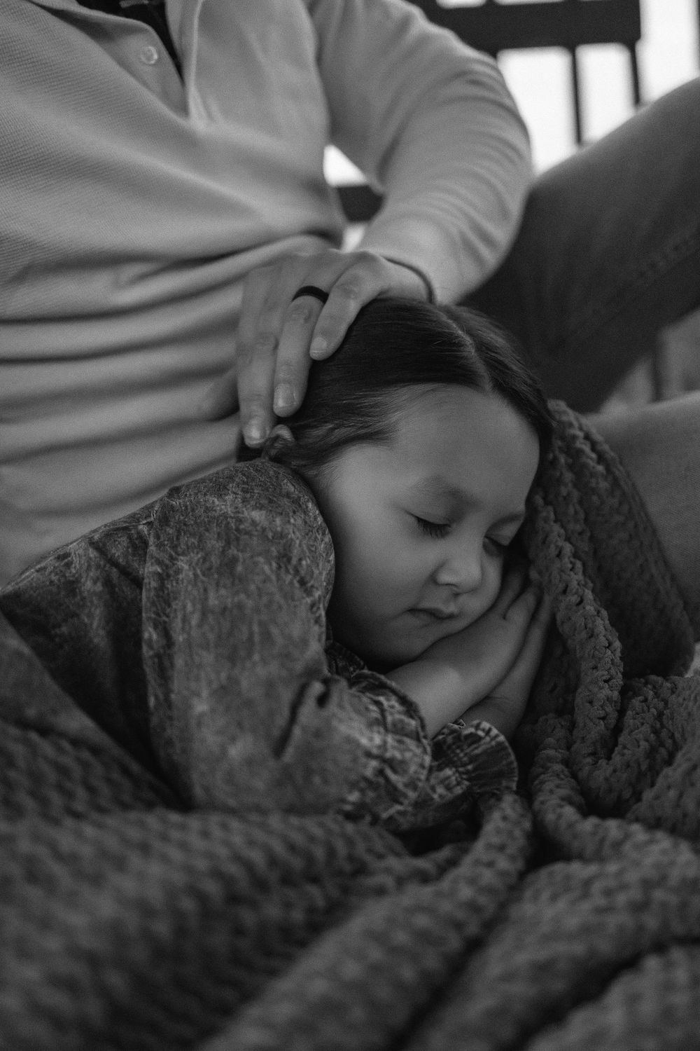 A black and white photo showing a 3 year old girl sleeping peacefully under a textured blanket with her Dad's hand placed on her head.