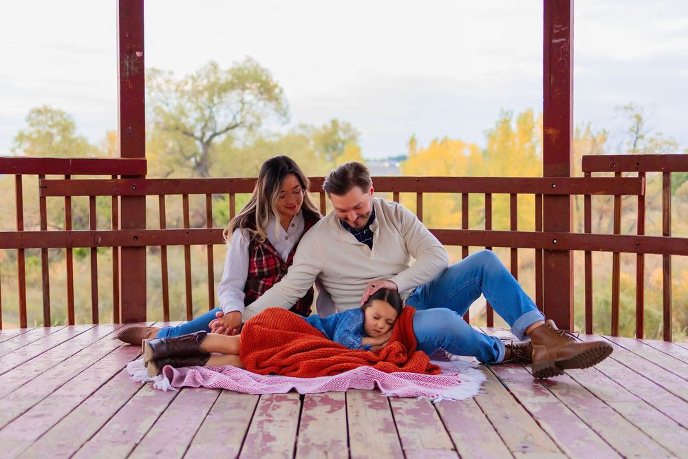 A family relaxes together on a blanket on a wooden deck during autumn with fall foliage in the background.