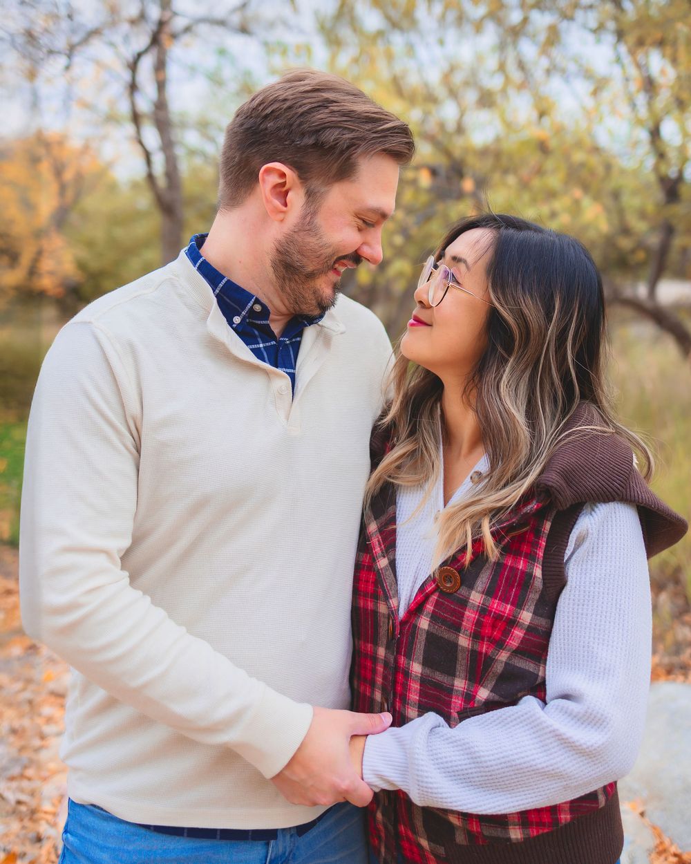 A couple in coordinating red plaid and cream sweater outfits embrace during an autumn outdoor photoshoot.