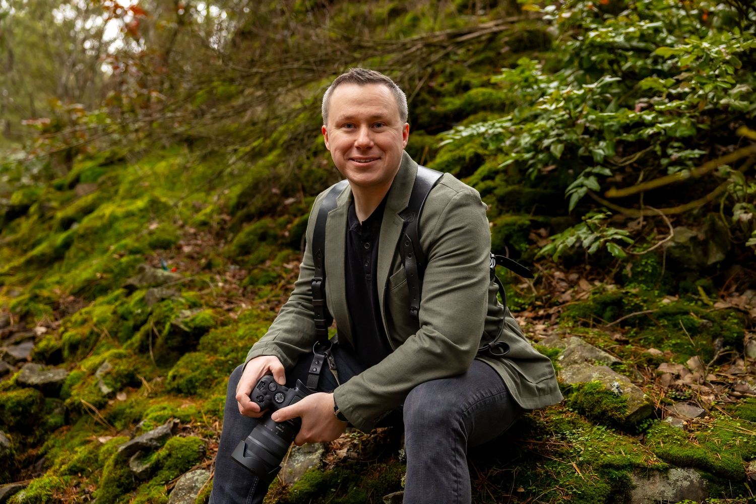 A person in a gray jacket sits on mossy rocks in a lush forest setting during autumn.