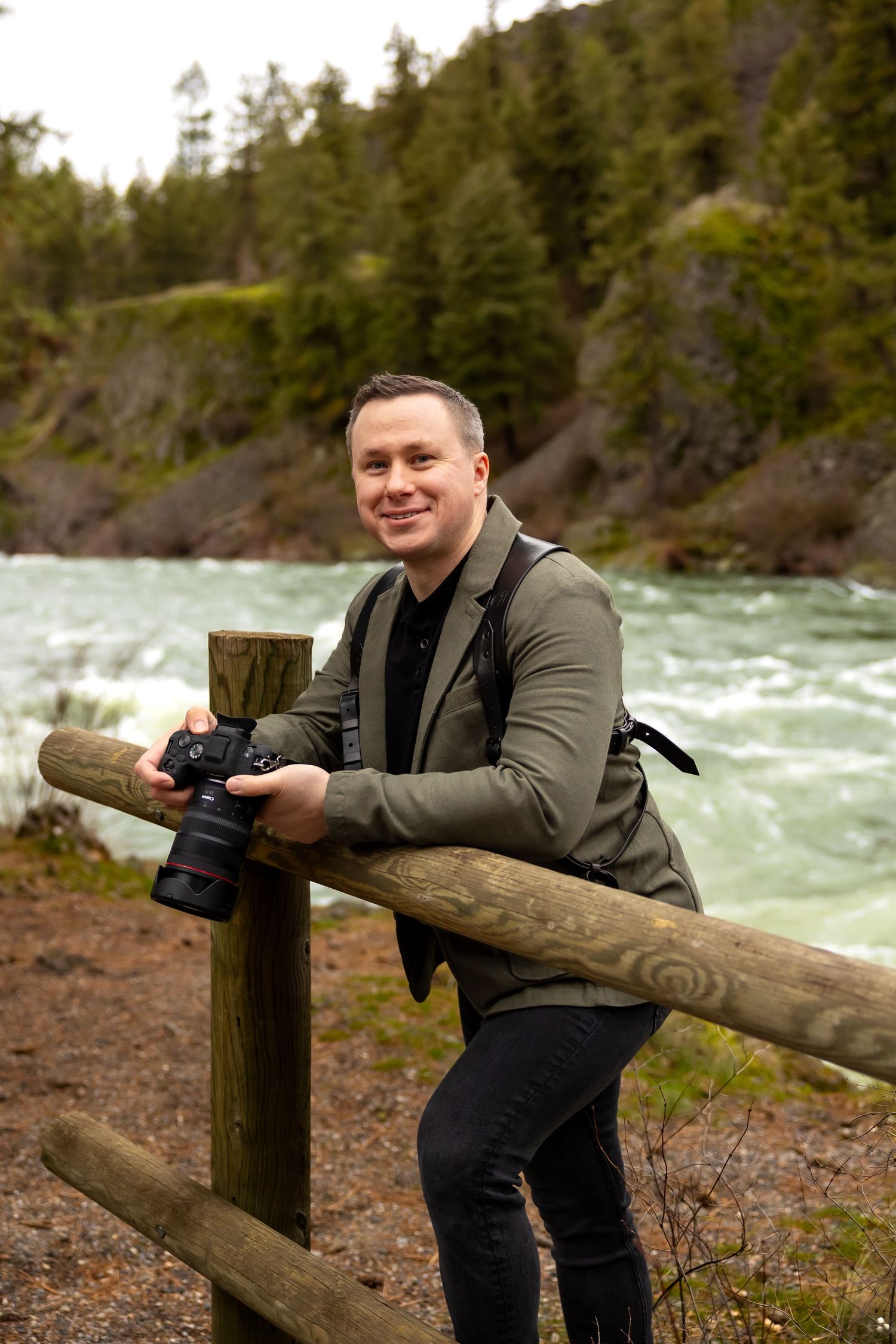 A photographer with camera equipment stands by a wooden railing near rushing river rapids in a forested area.