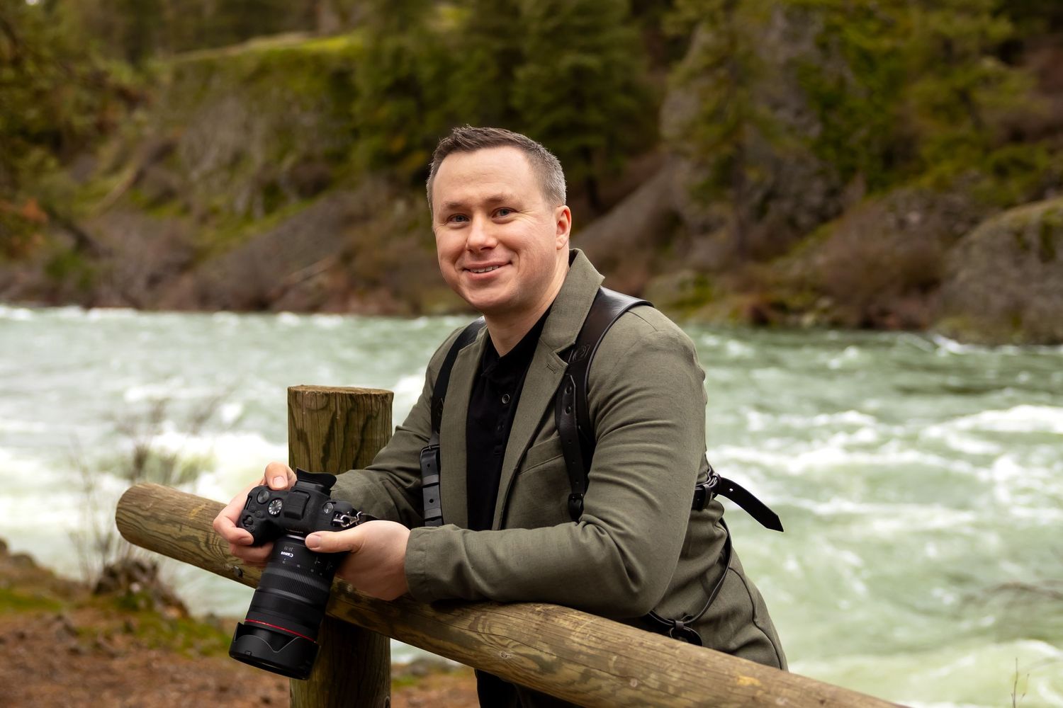A photographer with a camera stands by a rushing river in nature wearing a green jacket.
