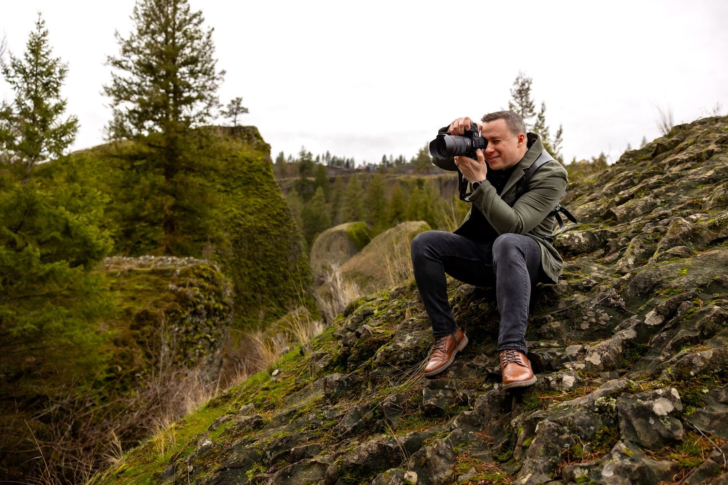A photographer crouches on rocky terrain with camera equipment while capturing nature shots in a mountainous setting.