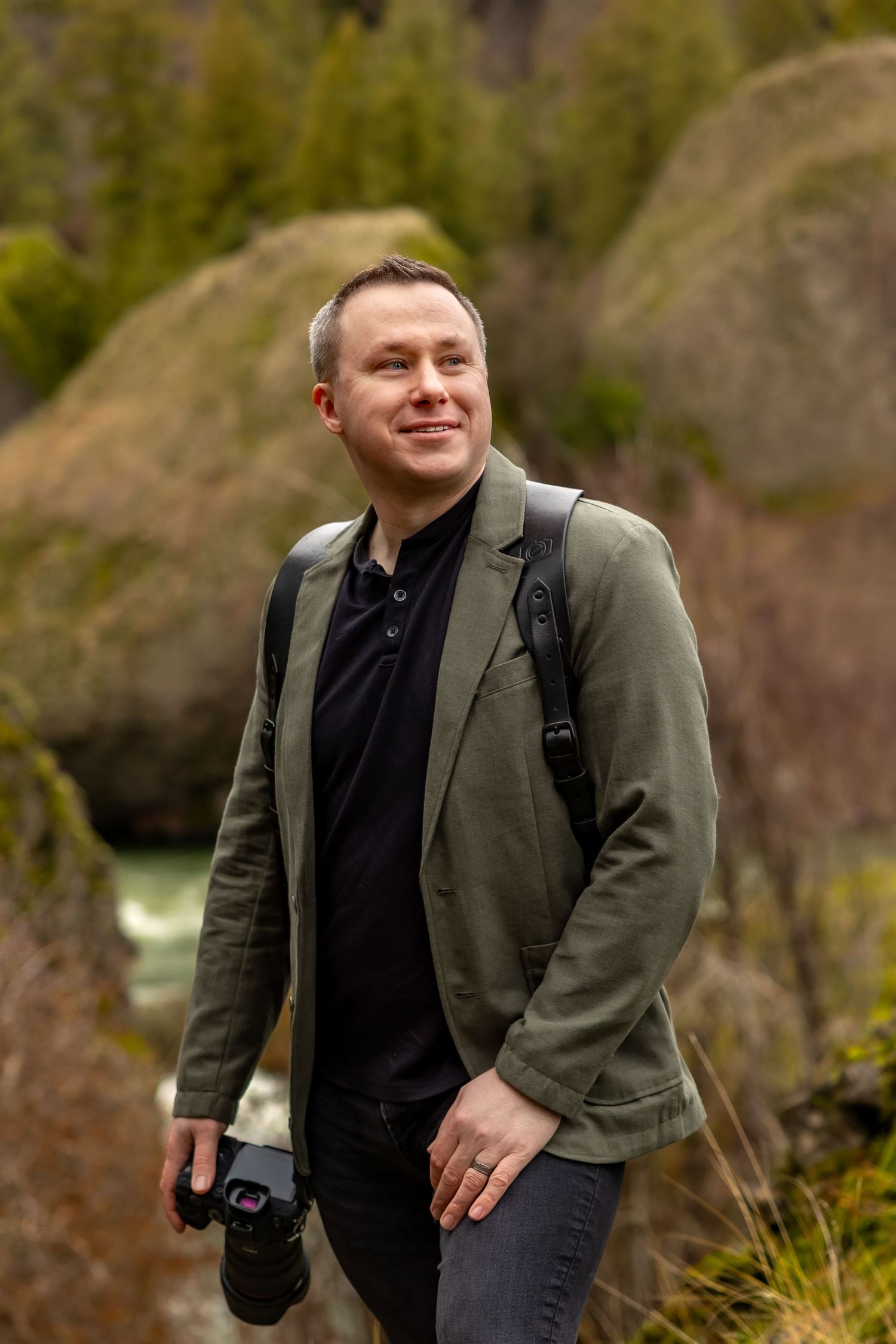A person wearing a dark blazer and black shirt poses outdoors against a natural backdrop with greenery.