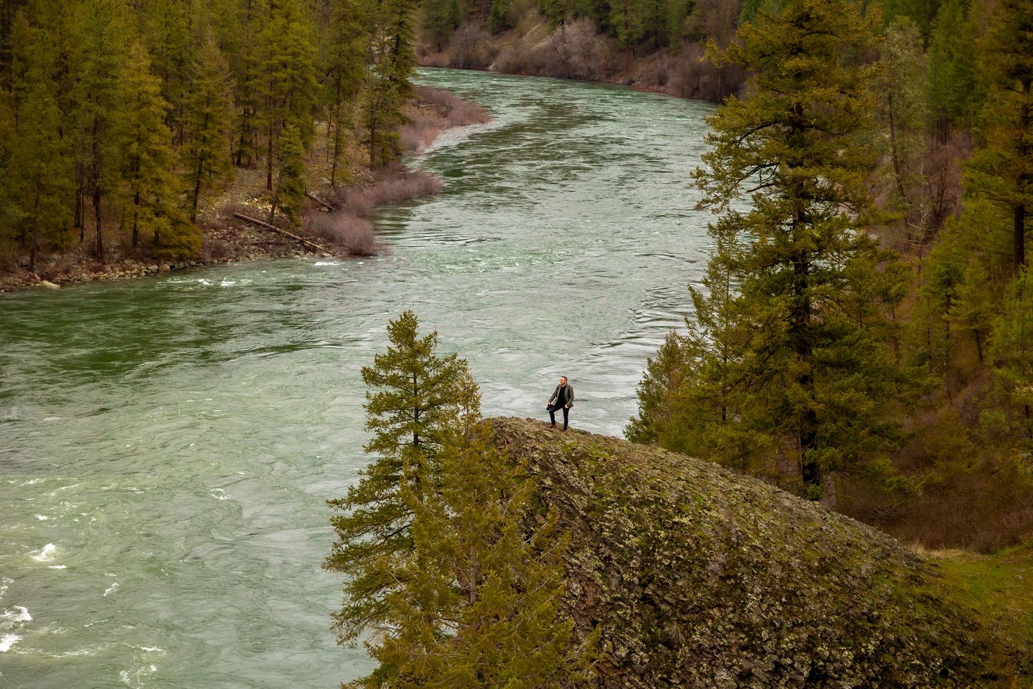 Turquoise river flows through dense evergreen forest with rocky cliffs and hiking trail along the riverbank.
