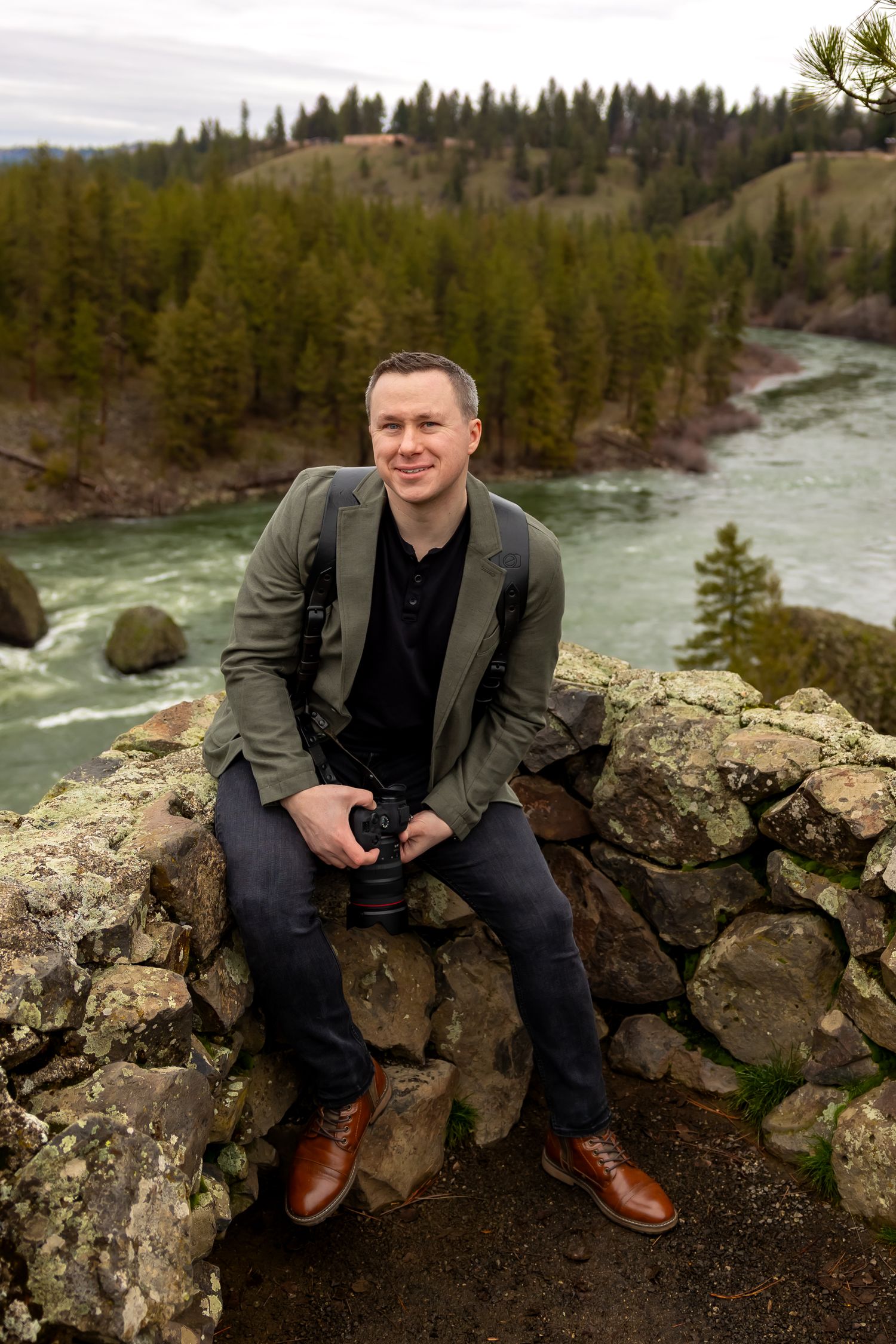 A person sits on rocky outcroppings beside a rushing river surrounded by evergreen forest in a scenic mountain landscape.