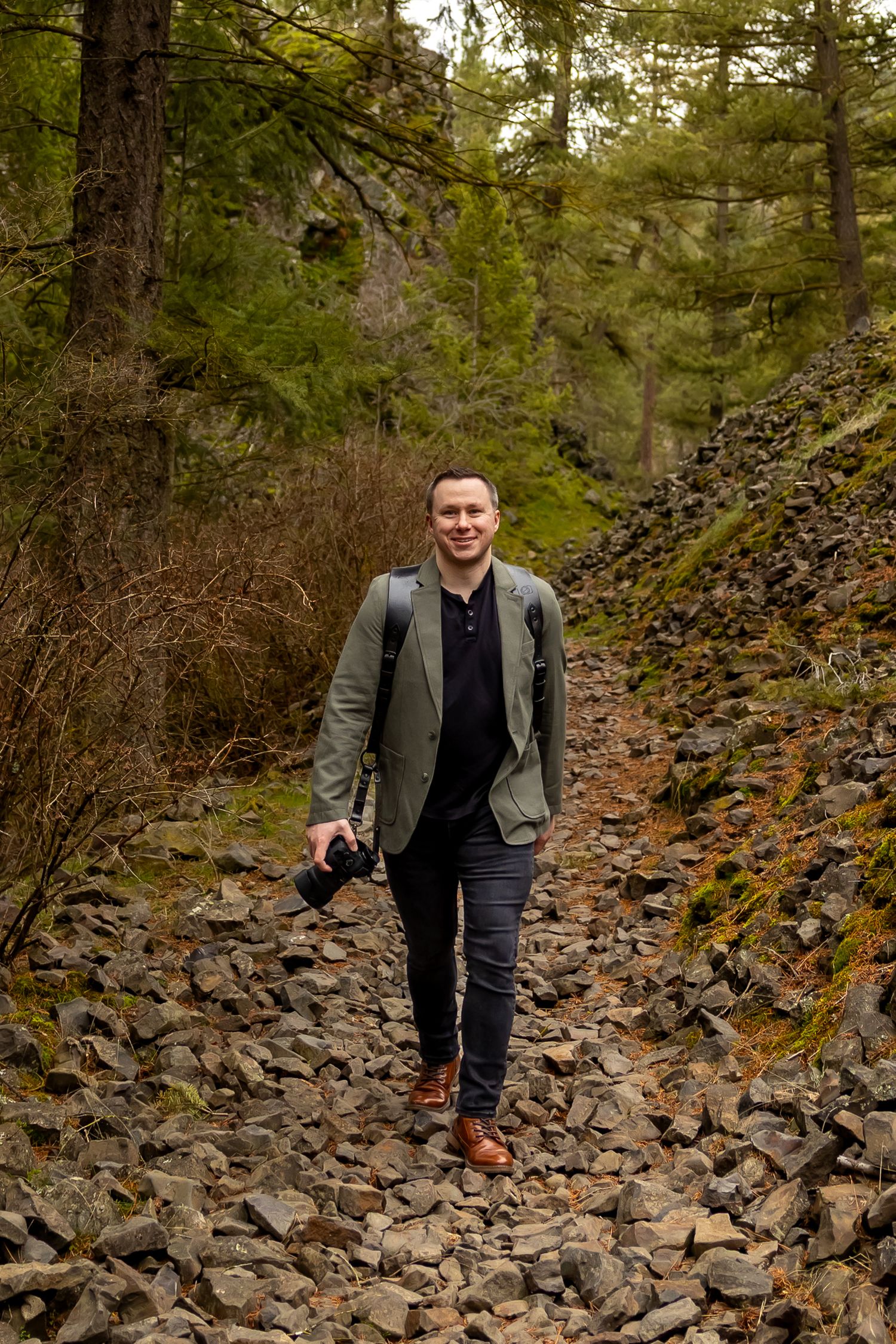 A person walks along a rocky forest trail surrounded by evergreen trees and autumn foliage.