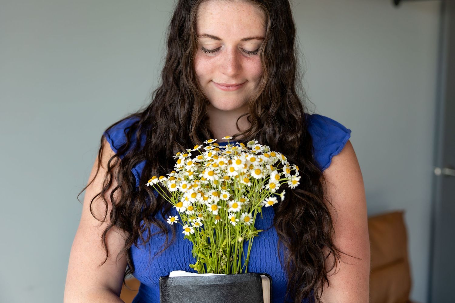 Woman in blue dress holds white daisies while smiling and looking down at fresh flowers.