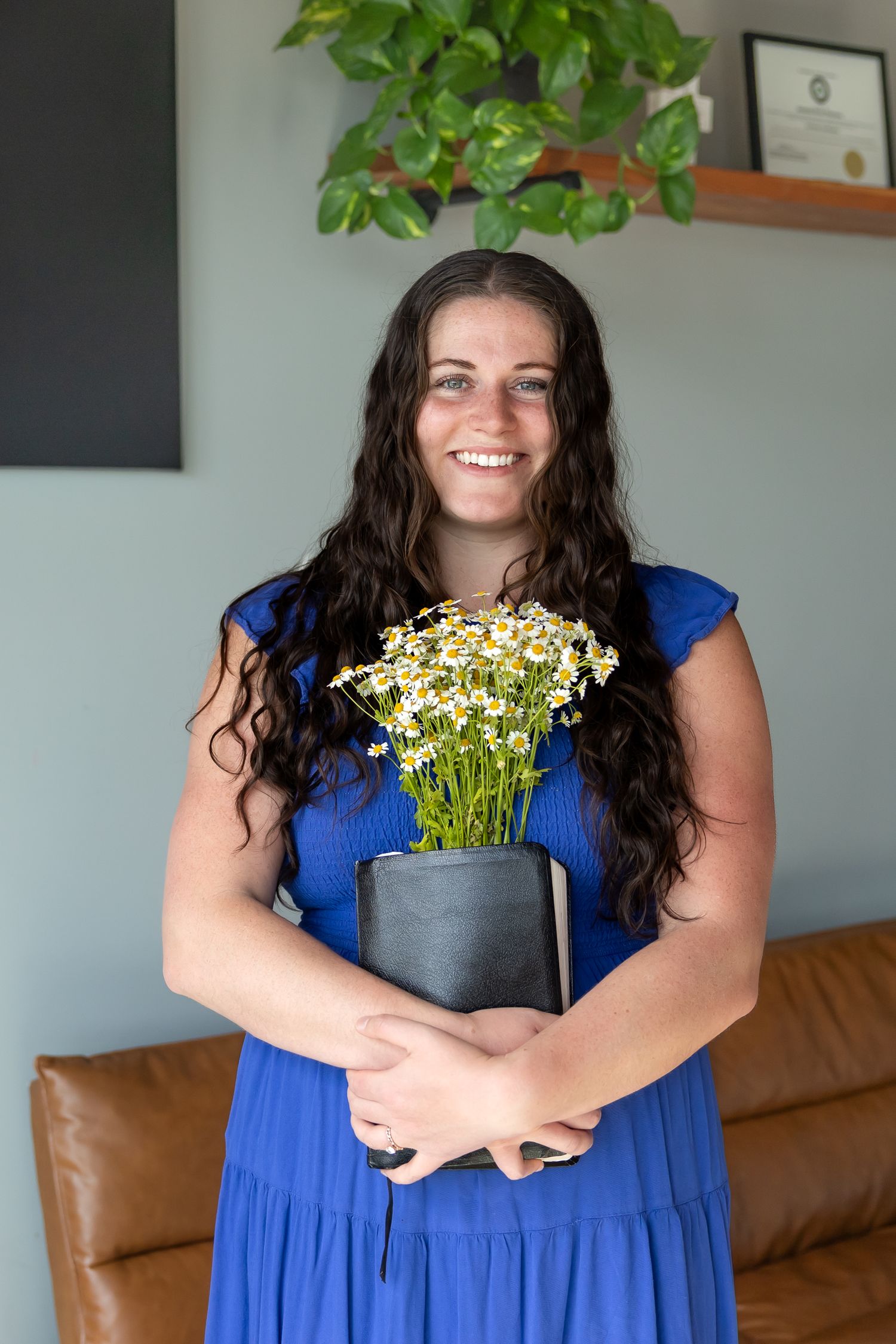 Someone wearing a royal blue dress holds a potted plant with small white flowers while seated on a leather couch.