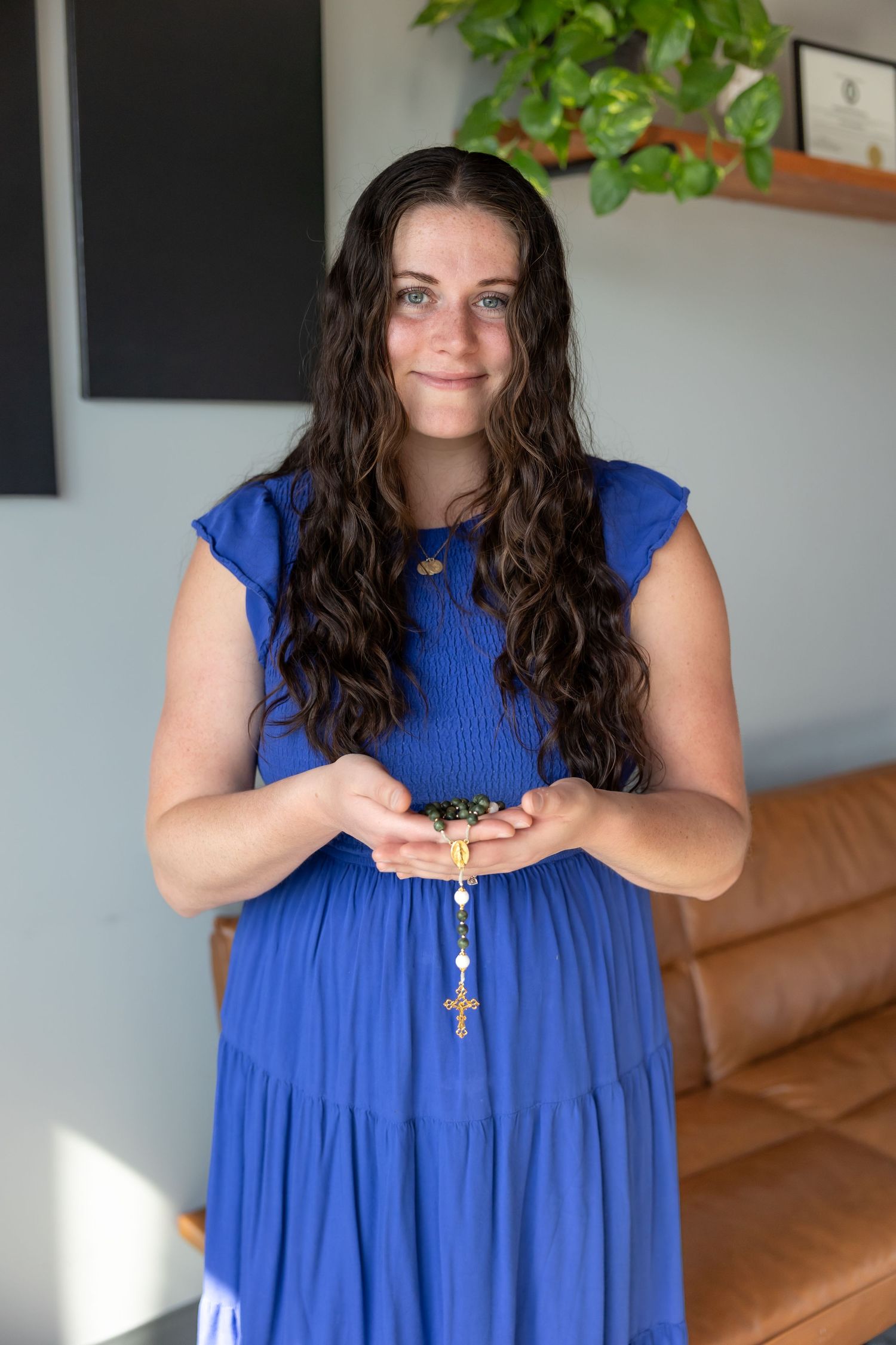 Woman wearing a vibrant royal blue sleeveless dress stands in a room with indoor plants and brown furniture.