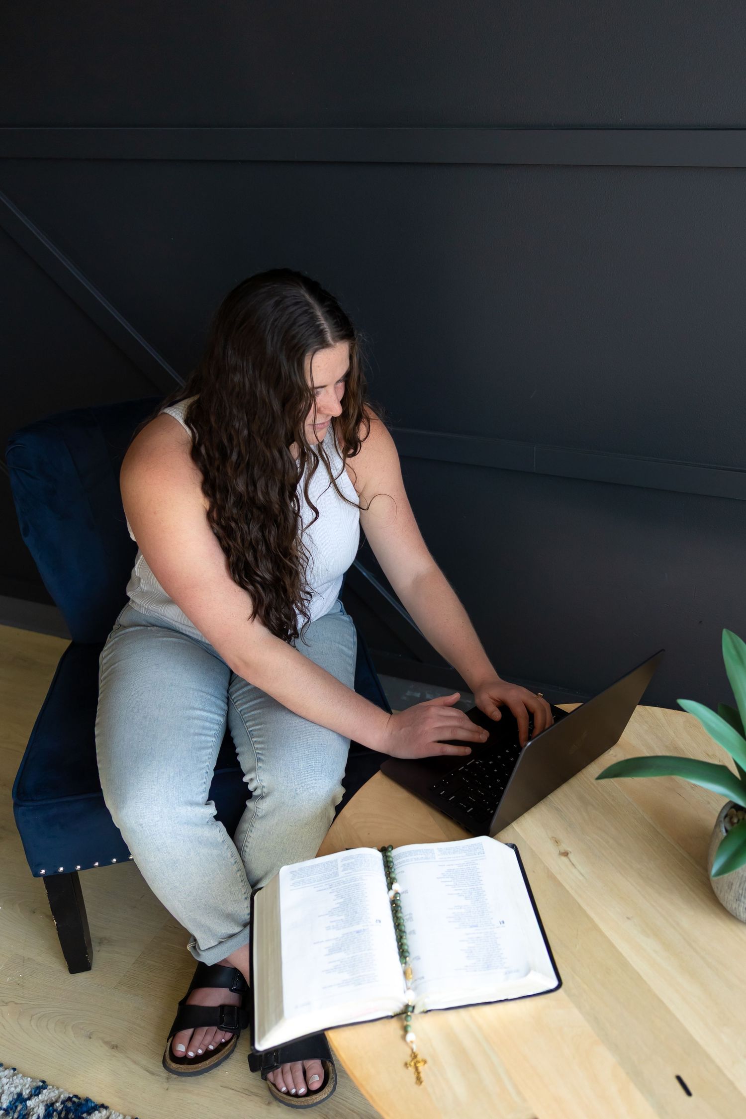 Person in light colored pants sits at wooden table reading from an open book with a cross necklace.