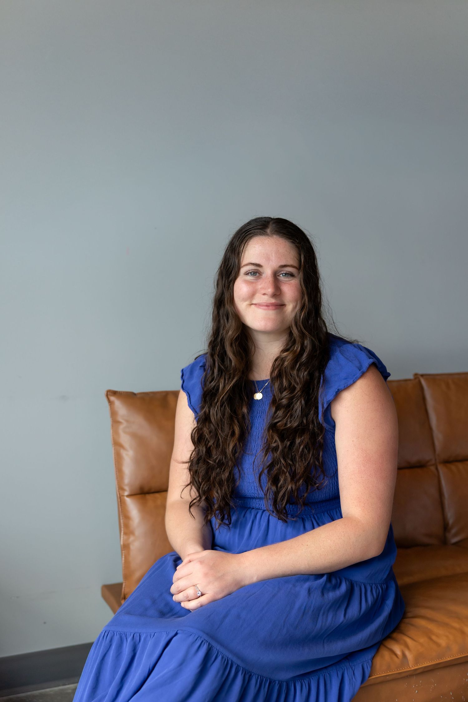 A person in a vibrant blue dress sits on a brown leather couch in a minimalist room setting.
