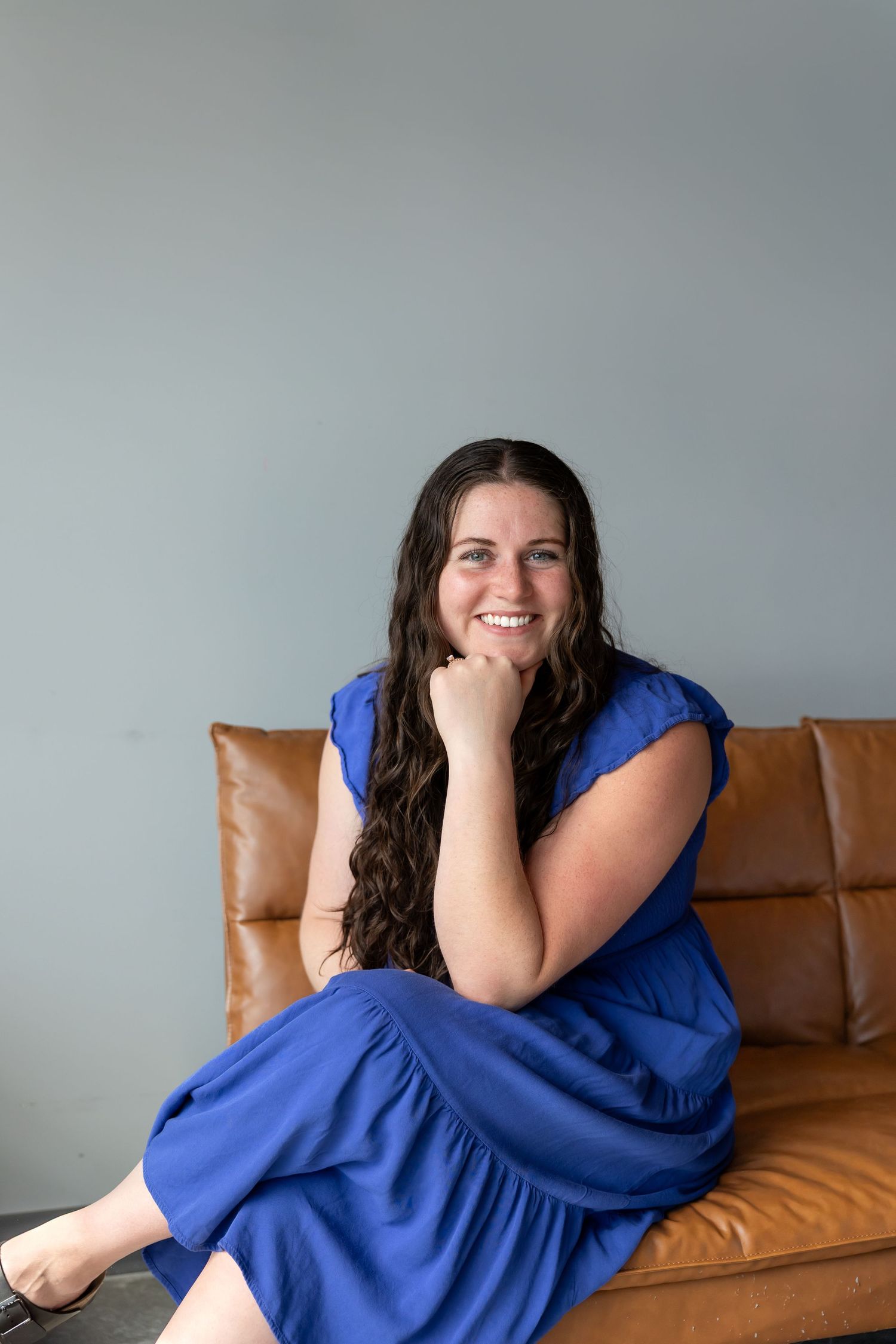 Woman in bright blue dress lounges on tan leather couch while smiling and laughing.