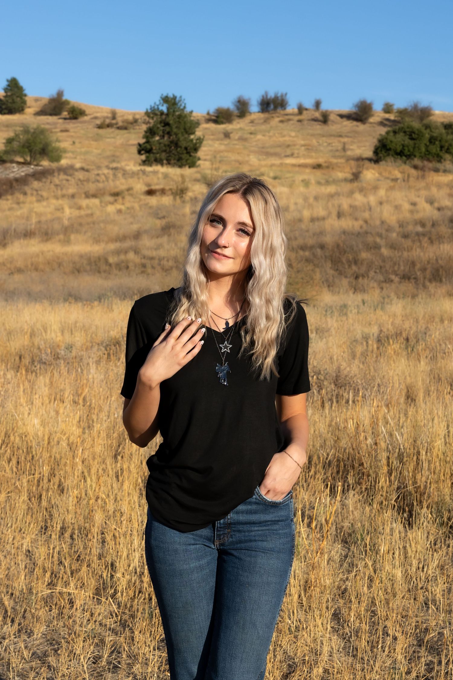 Person in black top and jeans poses against golden field on sunny day with blue sky in background.