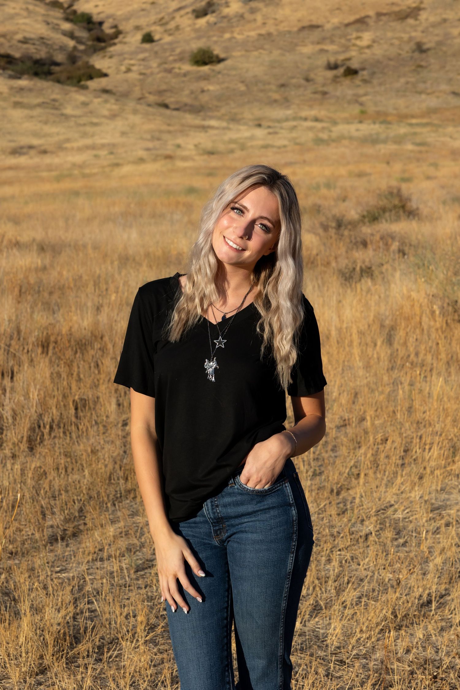 Person in black shirt and jeans poses playfully in a golden field with mountains in the background.