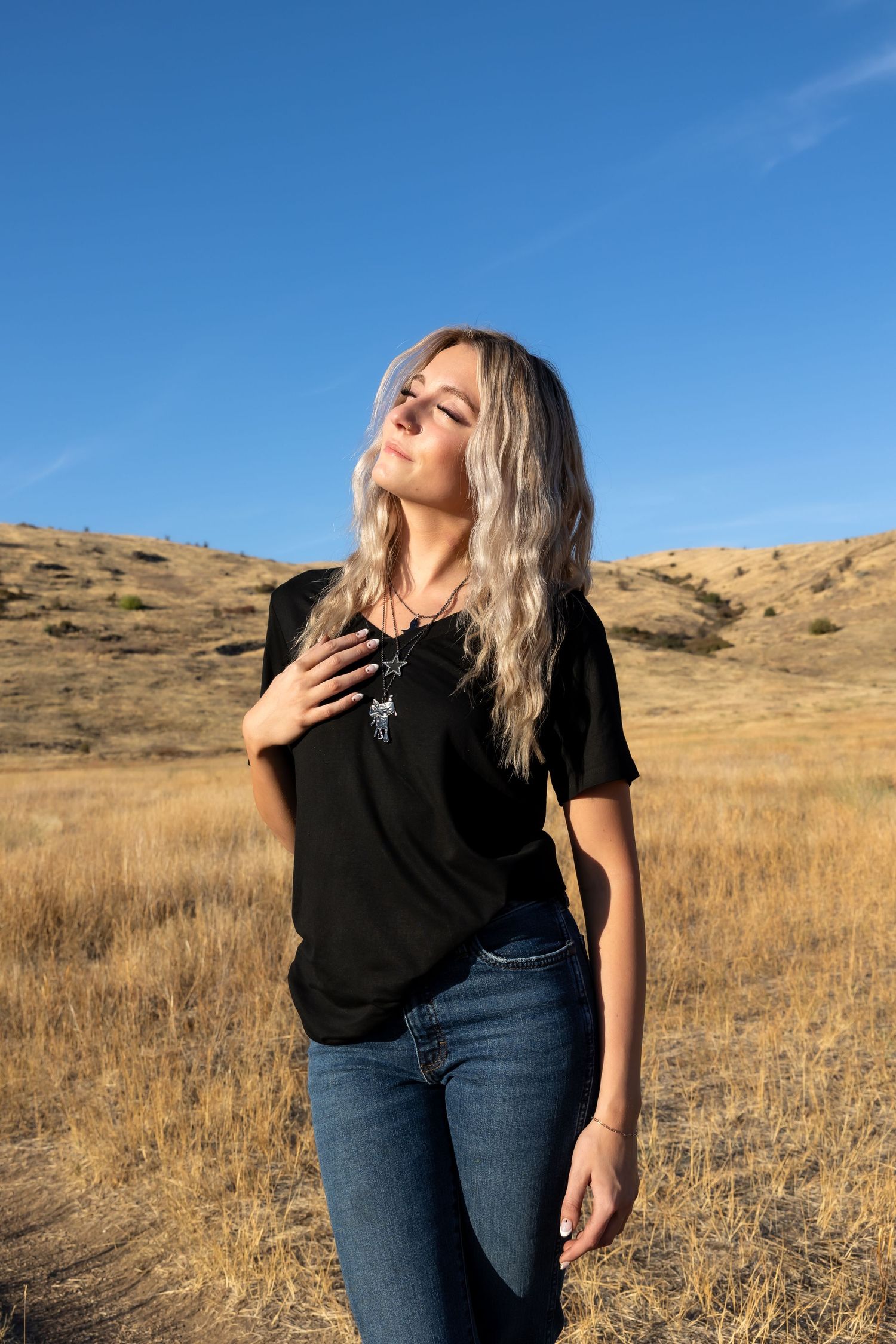 Person in black t-shirt poses against golden hillside and blue sky during outdoor photoshoot.