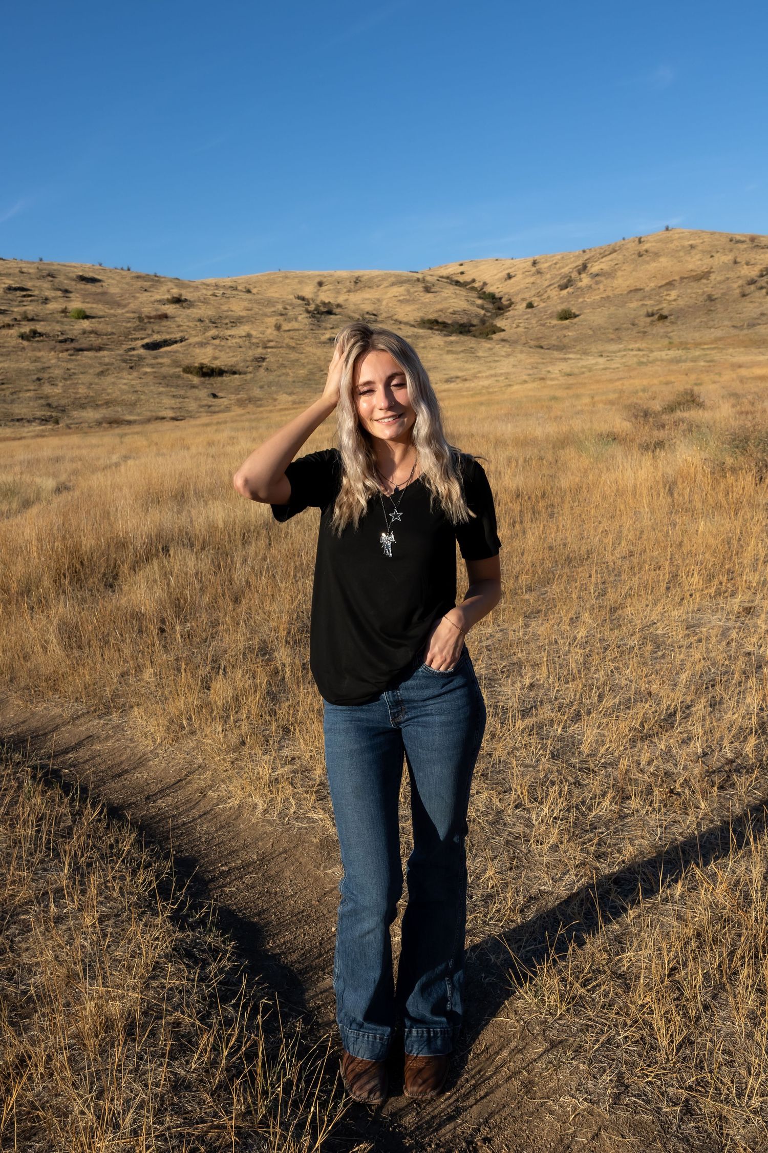 A person in black clothing poses against a backdrop of golden hills and blue sky on a sunny day.