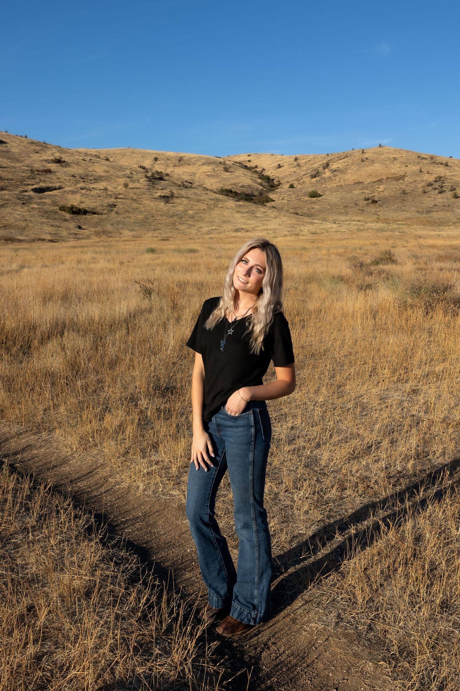Person in casual attire poses playfully in a golden field with rolling hills in the background during sunset.
