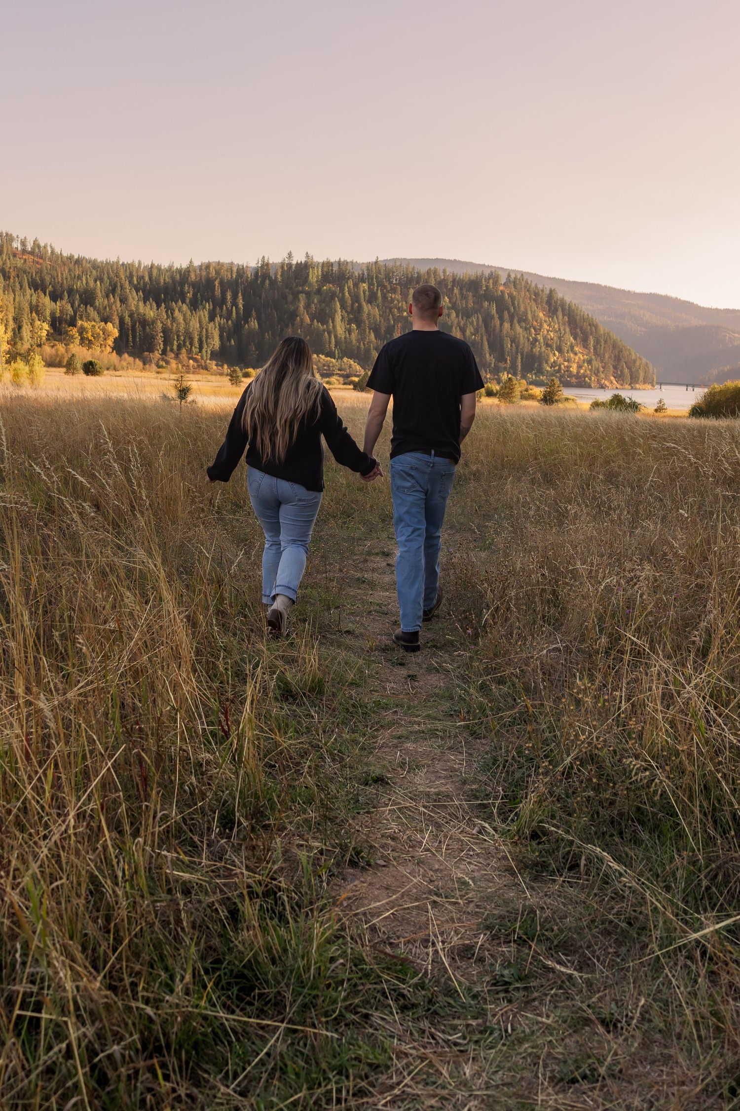A couple walking together on a scenic trail through tall grass at sunset with mountains and forest in the background.