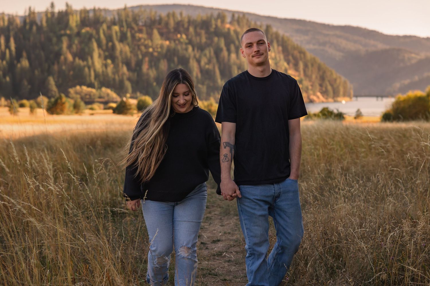A couple walks together in a golden field at sunset with mountains in the background.