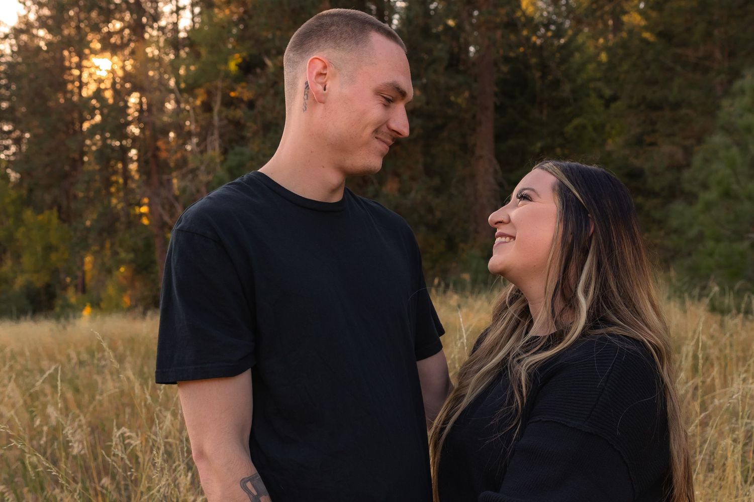 A couple embraces in a golden field at sunset with mountains and trees in the background.