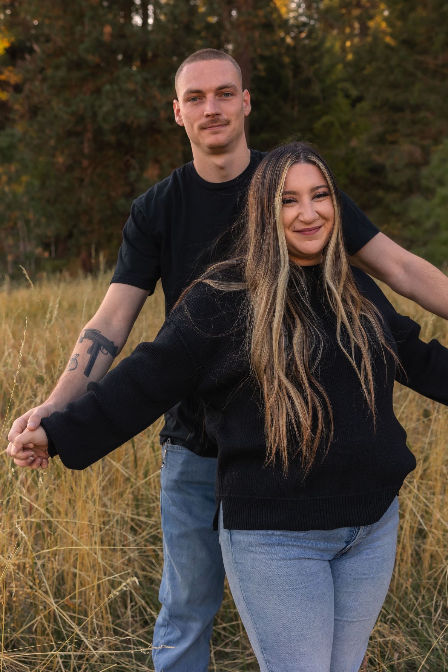 A couple in black sweaters and jeans playfully pose together in a golden field at sunset.