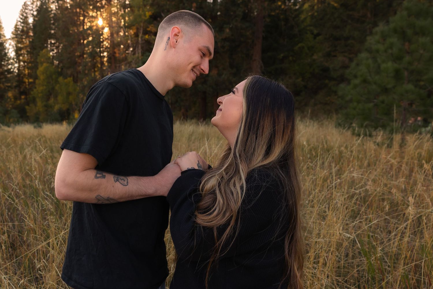 A couple shares a romantic moment in a golden field at sunset with tall trees in the background.