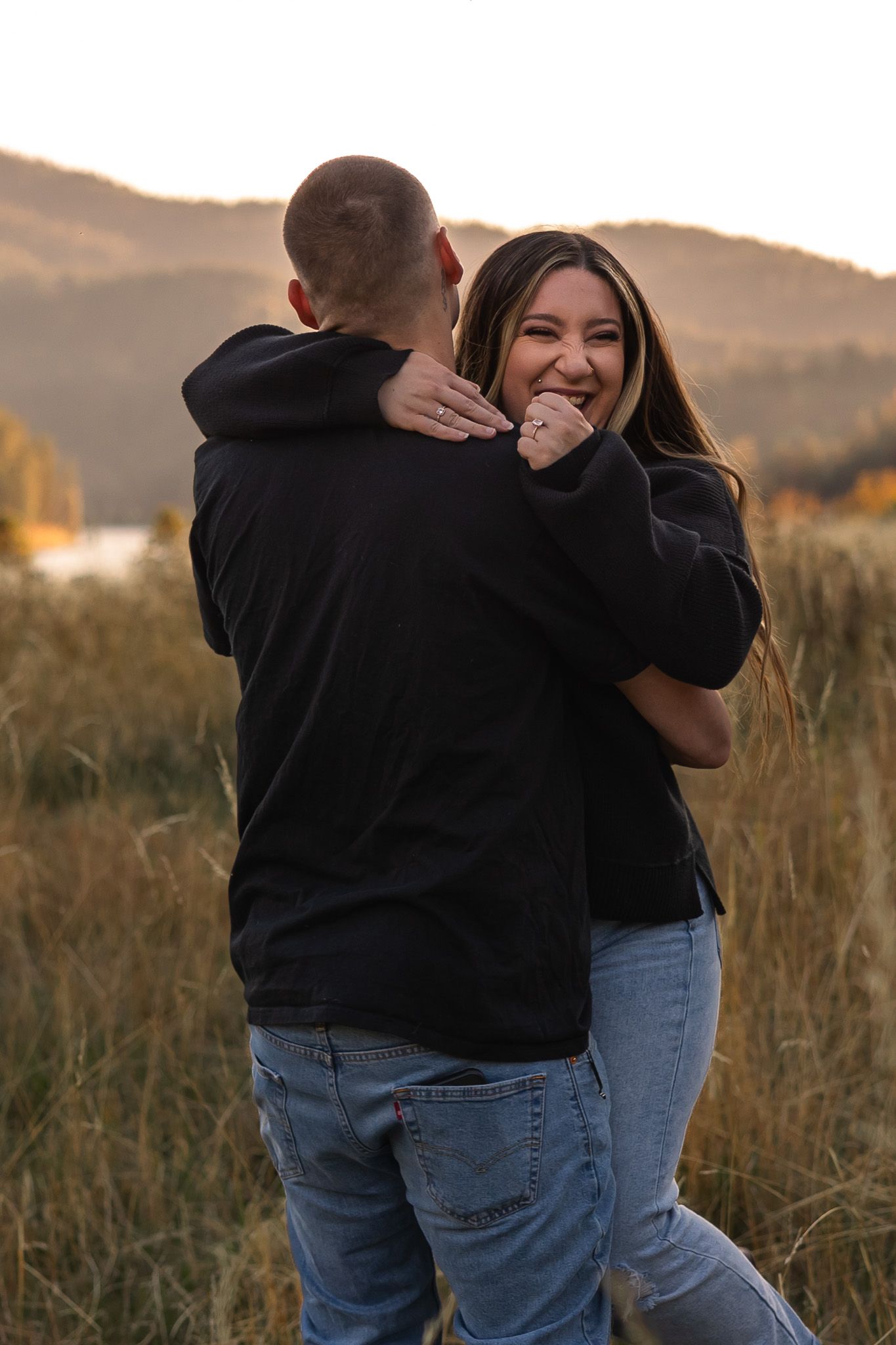 Two people embrace in a golden field at sunset with mountains in the background.